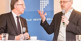 Zwei Männer in Anzügen sitzen an einem Tisch mit Mikrofonen und diskutieren. Einer gestikuliert mit der Hand, während der andere zuhört. Dahinter ist ein blauer Hintergrund mit weißem Text und einem Kartenmuster zu sehen. Auf dem Tisch stehen Wassergläser.