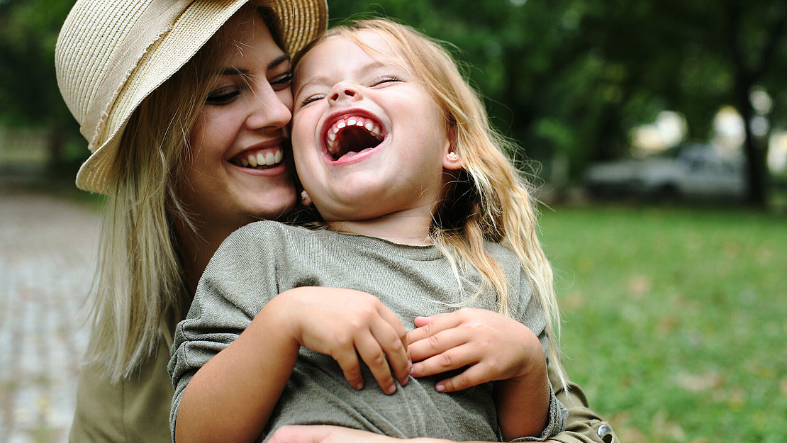 Eine Frau mit Hut umarmt im Freien ein lachendes junges Mädchen. Beide lächeln fröhlich, umgeben von Grün und einem unscharfen Hintergrund, was eine warme und fröhliche Atmosphäre schafft.