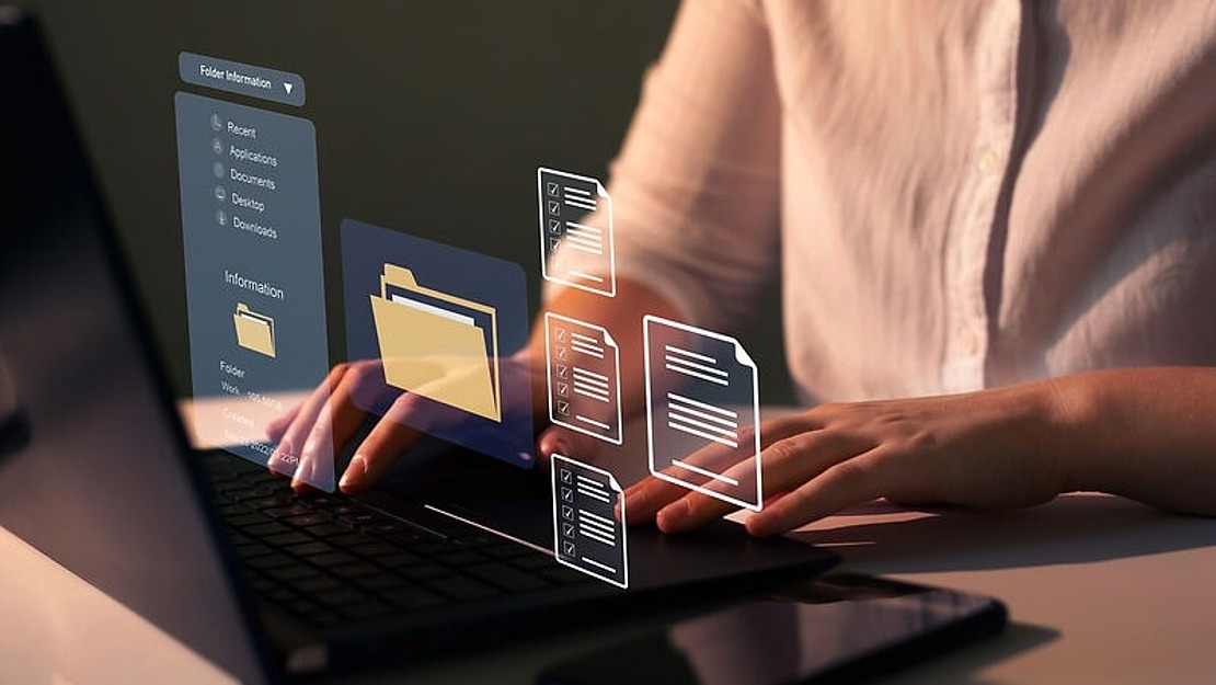 Person tippt auf einem Laptop, über dessen Tastatur eine Anzeige digitaler Ordner und Dokumente schwebt, die auf Datenverwaltung oder -organisation hindeutet. Die Szene ist schwach beleuchtet, wodurch die virtuelle Schnittstelle hervorgehoben wird.