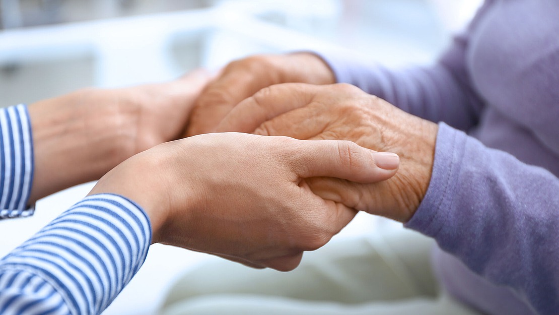 Nahaufnahme einer jüngeren Person, die sanft die Hände einer älteren Person hält, beide sitzend. Die ältere Person trägt einen lilafarbenen Pullover, die jüngere Person ein blau-weiß gestreiftes Hemd.