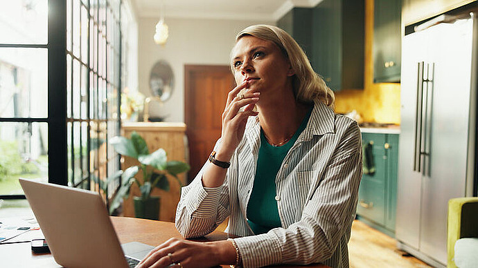 Eine Frau sitzt an einem Tisch in einer modernen Küche und arbeitet nachdenklich mit der Hand vor dem Mund an einem Laptop. Durch die großen Fenster neben ihr fällt Sonnenlicht ein.