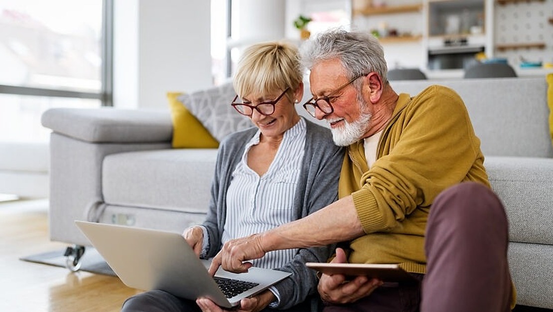 Älteres Paar sitzt in einem Wohnzimmer auf dem Boden, lächelt und schaut auf einen Laptop. Der Mann, der eine Brille und einen gelben Pullover trägt, hält ein Tablet in der Hand. Die Frau, die ebenfalls eine Brille trägt, zeigt auf den Laptop-Bildschirm. Im Hintergrund ist eine Couch zu sehen.