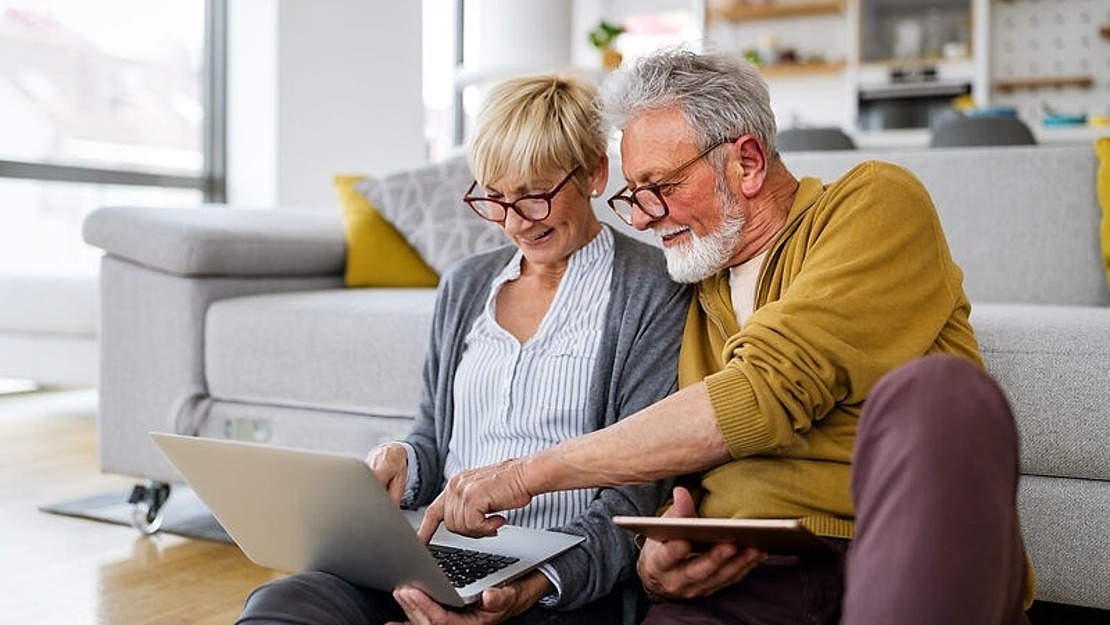Älteres Paar sitzt in einem Wohnzimmer auf dem Boden, lächelt und schaut auf einen Laptop. Der Mann, der eine Brille und einen gelben Pullover trägt, hält ein Tablet in der Hand. Die Frau, die ebenfalls eine Brille trägt, zeigt auf den Laptop-Bildschirm. Im Hintergrund ist eine Couch zu sehen.