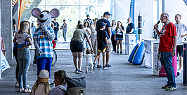 Eine Person in einem Maus-Maskottchen-Kostüm unterhält sich mit Kindern und Erwachsenen bei einer Indoor-Veranstaltung. Menschen stehen in Gruppen, einige unterhalten sich, während eine andere Person in ein Mikrofon spricht. Banner und Veranstaltungsstände sind zu sehen.