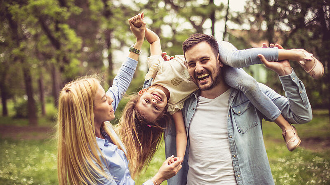 Ein lächelnder Mann hält ein lachendes junges Mädchen kopfüber, während eine Frau daneben steht. Alle genießen die gemeinsame Zeit im Freien in einem Park mit grünen Bäumen im Hintergrund.