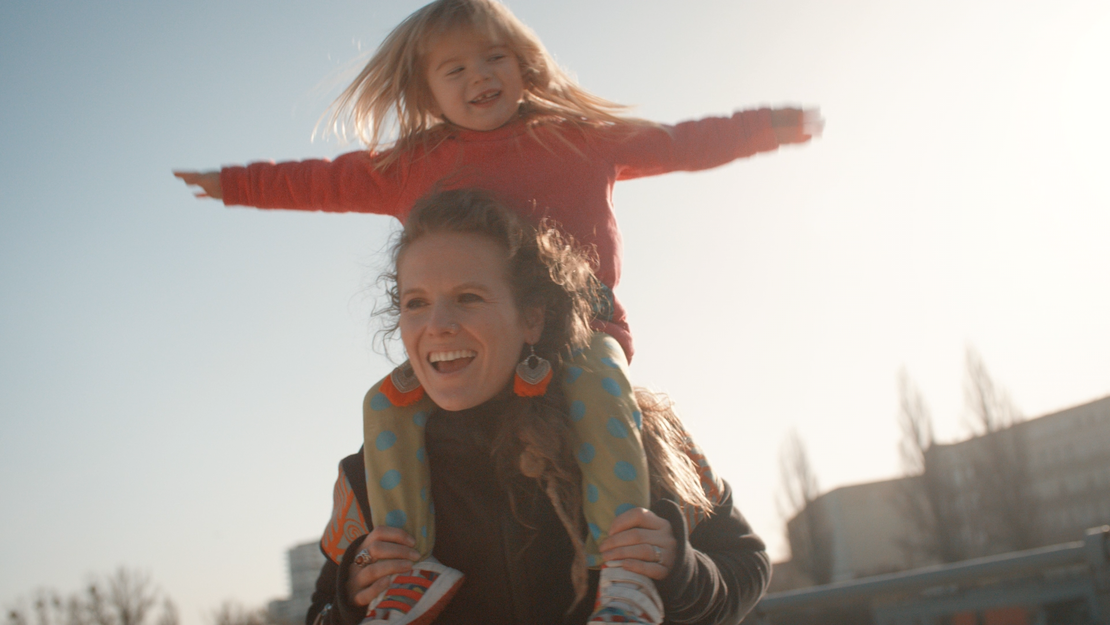 Eine lächelnde Frau trägt ein fröhliches junges Mädchen auf ihren Schultern im Freien. Das Mädchen streckt die Arme weit aus, beide genießen den sonnigen Tag. Im unscharfen Hintergrund sind Gebäude und Bäume zu sehen.