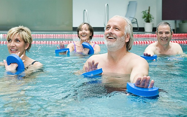Vier Erwachsene trainieren in einem Schwimmbecken mit blauen Schaumstoffpaddeln. Sie lächeln und scheinen an einem Gruppentraining teilzunehmen. Im Hintergrund sind Schwimmbahnen und Liegestühle am Beckenrand zu sehen.