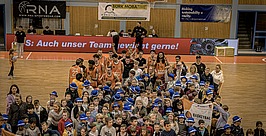 Eine große Gruppe von Kindern posiert mit Basketballspielern auf einem Hallenplatz. Viele Kinder tragen blaue Mützen und halten Basketballartikel in der Hand. Die Spieler tragen orangefarbene Trikots. Im Hintergrund sind Banner und Werbung zu sehen.