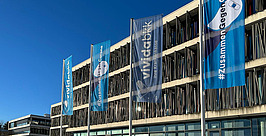 Ein modernes Gebäude mit großen Fenstern steht unter einem strahlend blauen Himmel. An vier hohen Fahnenmasten hängen blaue Banner, auf einem steht „#ZusammenGegenCorona“ mit der Abbildung einer Impfspritze. Schnee bedeckt den Boden.