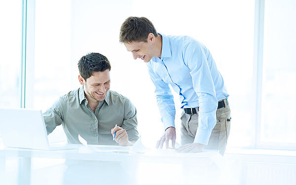 Zwei Männer sitzen an einem Schreibtisch in einem hellen Büro. Einer steht und zeigt auf Dokumente auf dem Tisch, während der andere sitzt, lächelt und schreibt. In der Nähe steht ein geöffneter Laptop, und große Fenster sorgen für natürliches Licht.