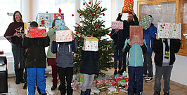 Eine Gruppe von Kindern und zwei Erwachsenen stehen drinnen um einen geschmückten Weihnachtsbaum und halten sich eingepackte Geschenke vors Gesicht. Weitere Geschenke liegen verstreut unter dem Baum. Alle tragen Winterkleidung.