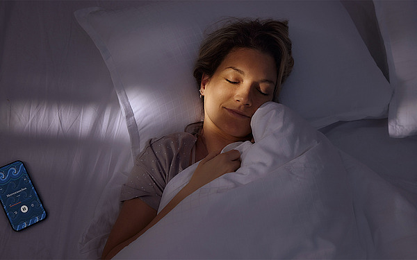 Eine Person liegt friedlich schlafend im Bett und lächelt sanft. Neben ihr liegt ein Smartphone mit blauem Bildschirm, auf dem eine Schlaf-App angezeigt wird. Der Raum ist schwach beleuchtet, was eine ruhige und heitere Atmosphäre schafft.