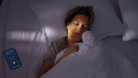 Eine Person liegt friedlich schlafend im Bett und lächelt sanft. Neben ihr liegt ein Smartphone mit blauem Bildschirm, auf dem eine Schlaf-App angezeigt wird. Der Raum ist schwach beleuchtet, was eine ruhige und heitere Atmosphäre schafft.