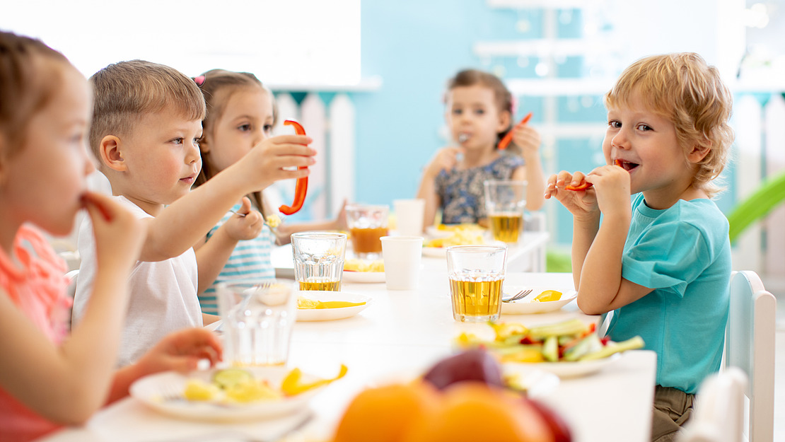 Kinder sitzen an einem Tisch in einem hellen Raum und genießen eine Mahlzeit. Obst, Gemüse und Getränke stehen auf dem Tisch. Die Kinder lächeln und unterhalten sich und schaffen so eine fröhliche Atmosphäre. Im Hintergrund ist farbenfrohes Dekor zu sehen.