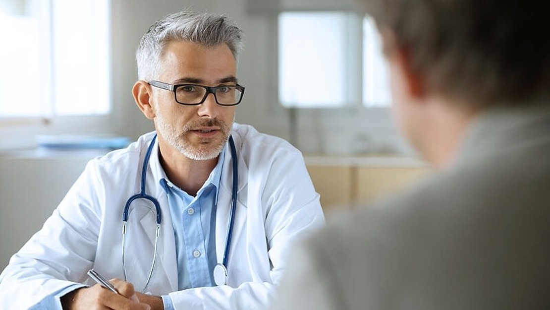 Ein Arzt mit grauem Haar, Brille und Bart sitzt an einem Schreibtisch, hält einen Stift in der Hand und spricht mit einer Person, die der Kamera den Rücken zuwendet. Der Arzt trägt einen weißen Kittel und ein Stethoskop um den Hals.