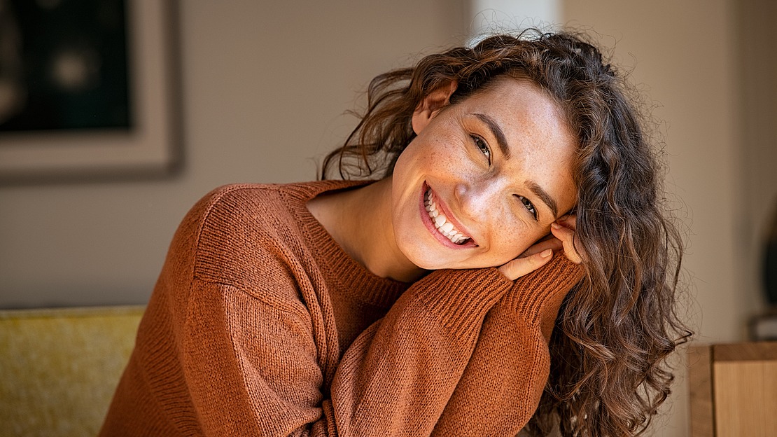 Eine Person mit lockigem Haar und einem braunen Pullover lächelt und stützt den Kopf auf die Hände. Sie sitzt im Innenbereich, im Hintergrund stehen ein gerahmtes Bild und einige Möbel.