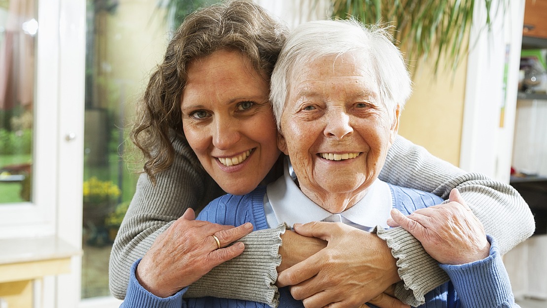 Eine lächelnde ältere Frau in einem blauen Pullover wird von hinten von einer jüngeren Frau mit lockigem Haar umarmt. Beide sehen glücklich aus und stehen in einem hell erleuchteten Raum mit Pflanzen.