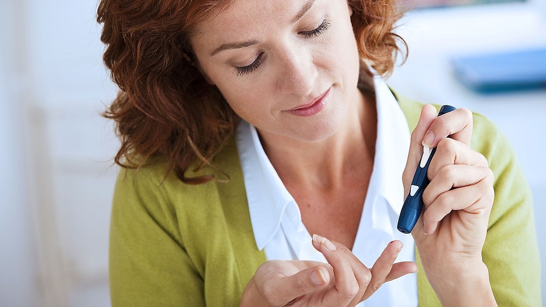 Eine Frau mit roten Locken und grüner Strickjacke benutzt ein Blutzuckermessgerät, um ihren Blutzucker durch Stechen in den Finger zu messen.