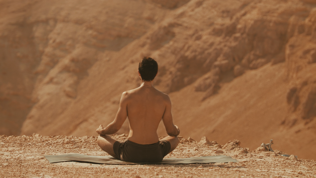 Eine Person ohne Hemd sitzt im Schneidersitz auf einer Matte in einer Wüstenlandschaft und meditiert mit dem Rücken zur Kamera inmitten eines felsigen, sandigen Geländes.
