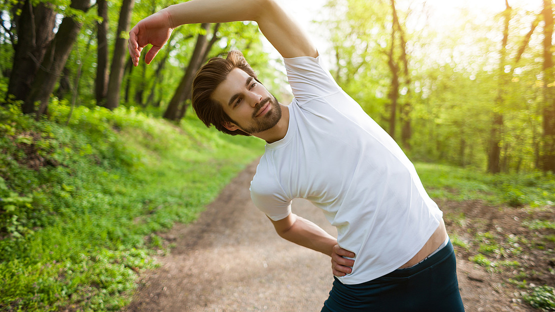 Ein junger Mann in einem weißen T-Shirt streckt seine Seite im Freien auf einem Waldweg, umgeben von grünen Bäumen und Sonnenlicht.