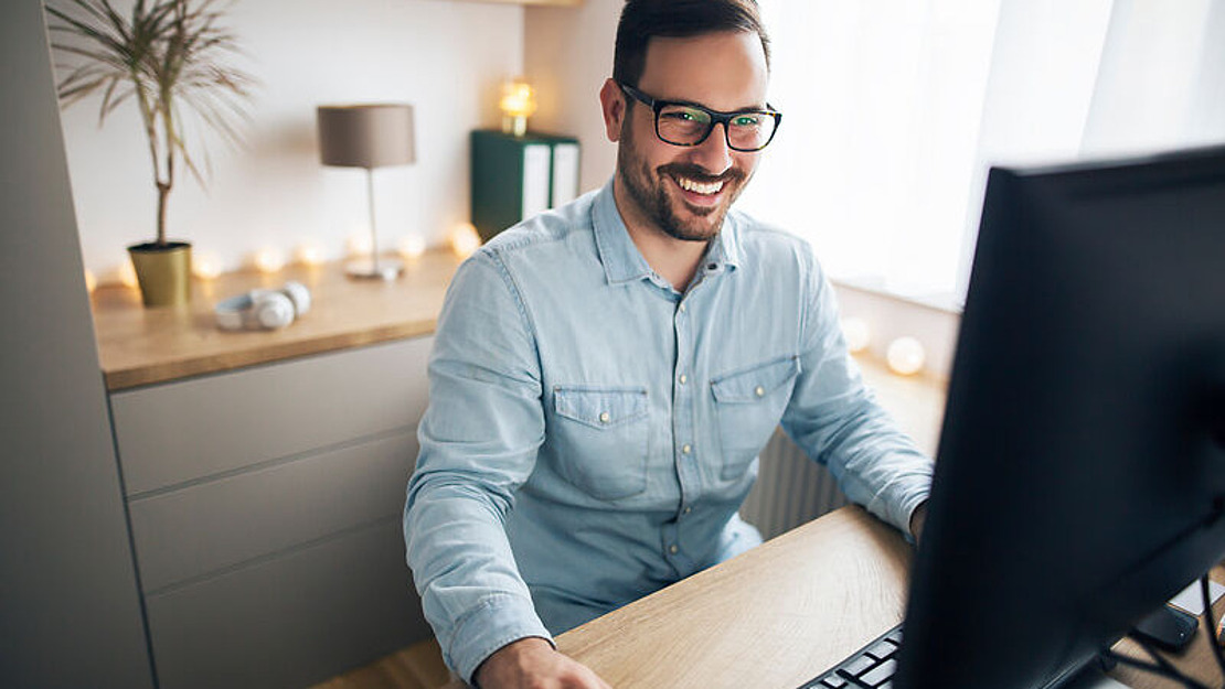 Ein lächelnder Mann mit Brille sitzt an einem hölzernen Schreibtisch und arbeitet an einem Computer in einem gemütlichen, gut beleuchteten Heimbüro mit dekorativen Lampen, Regalen und einer Topfpflanze im Hintergrund.