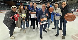 Eine Gruppe von neun Personen steht auf einer Eisbahn und lächelt in die Kamera. Einige halten Sportgeräte in den Händen, darunter Boxhandschuhe, einen Fußball und einen Basketball. Zwei Männer vor ihnen halten Veranstaltungsplakate. Hinter ihnen sind die Sitzplätze der Arena zu sehen.