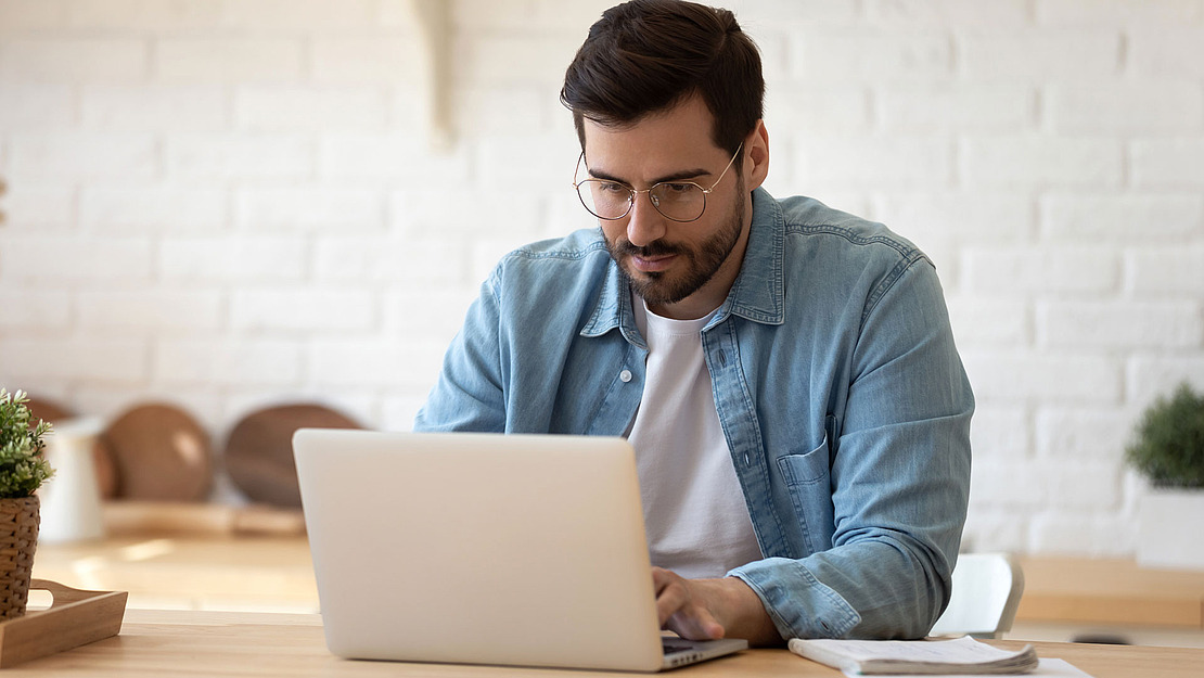 Ein Mann mit Brille und hellem Jeanshemd sitzt an einem Tisch und arbeitet konzentriert an seinem Laptop. Im Hintergrund ist ein heller Raum mit einer weißen Ziegelwand und minimalistischem Dekor zu sehen.