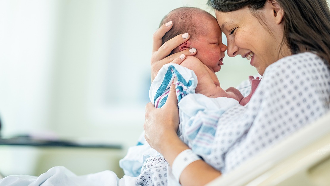 Eine Mutter im Krankenhaushemd wiegt liebevoll ihr Neugeborenes und berührt sanft die Stirn. Sie sitzt auf einem Krankenhausbett. Beide wirken friedlich und fröhlich. Der Raum ist von natürlichem Licht durchflutet.