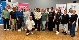 Eine Gruppe von 20 Personen, hauptsächlich Frauen, steht lächelnd für ein Gruppenfoto in einem Gebäude auf einem Holzboden. Ein Mann kniet davor. Im Hintergrund sind bunte Banner mit deutscher Aufschrift zu sehen.