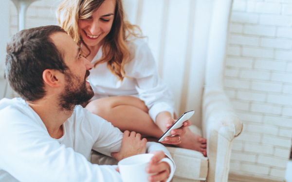 Ein Mann und eine Frau sitzen in einem gemütlichen, hellen Raum zusammen. Der Mann hält eine weiße Tasse in der Hand und lächelt die Frau an, die auf einem Stuhl sitzt, zurücklächelt und ein Smartphone in der Hand hält.