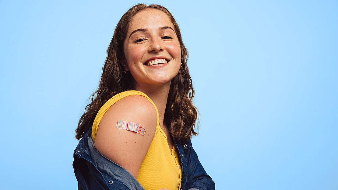 Eine lächelnde junge Frau mit langen braunen Haaren trägt ein gelbes Top und ein Jeanshemd. Auf ihrem Oberarm ist vor blauem Hintergrund ein bunter Verband zu sehen.