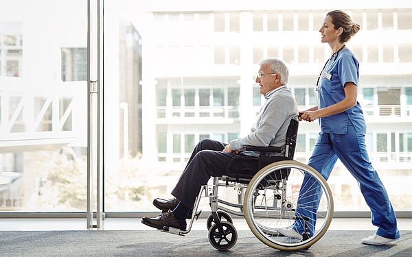 Eine Krankenschwester im blauen Kittel schiebt einen älteren Mann im Rollstuhl einen Flur mit großen Fenstern entlang. Die Szene wirkt hell und luftig, das Sonnenlicht fällt durch das Glas.