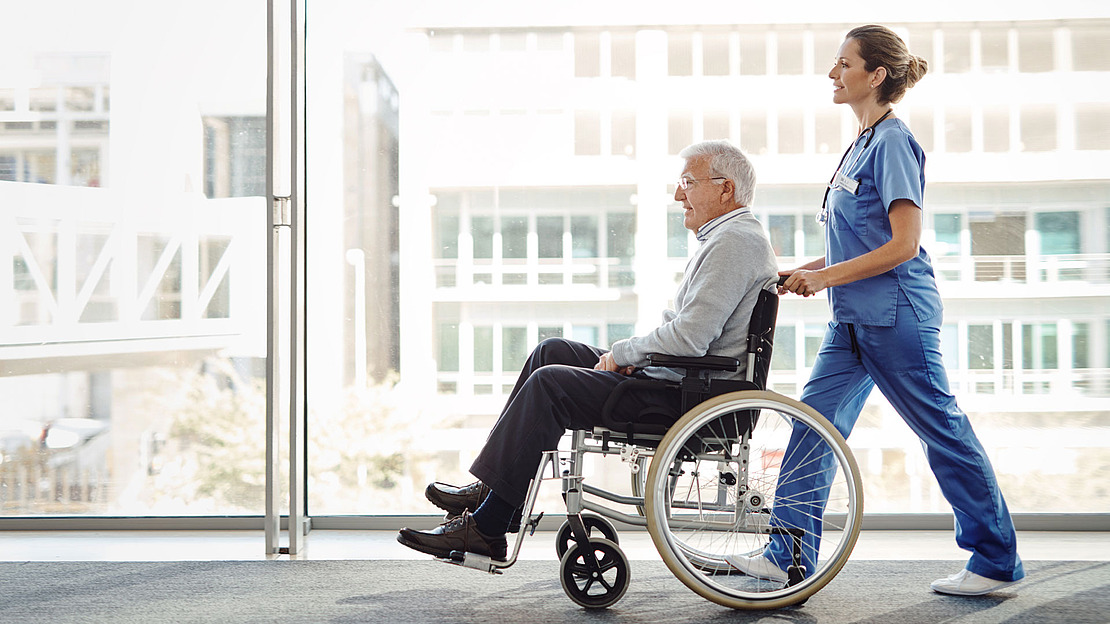 Eine Krankenschwester im blauen Kittel schiebt einen älteren Mann im Rollstuhl einen Flur mit großen Fenstern entlang. Die Szene wirkt hell und luftig, das Sonnenlicht fällt durch das Glas.