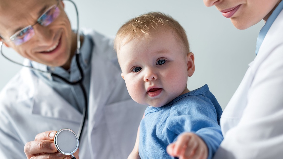Ein Baby in einem blauen Hemd sitzt auf dem Schoß eines Erwachsenen, während ein lächelnder Arzt in weißem Kittel und mit Brille ein Stethoskop in der Hand hält und sich darauf vorbereitet, die Brust des Babys bei einer medizinischen Untersuchung abzuhören.