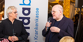 Zwei Personen stehen bei einer Veranstaltung in einem Gebäude und sprechen in Mikrofone. Im Hintergrund ist ein großes vertikales Schild mit blauer Schrift und dem Wort „Krankenkasse“ zu sehen. Die Frau hört zu, während der Mann gestikuliert.
