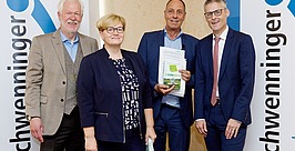 Vier professionell gekleidete Personen stehen lächelnd zwischen zwei Schwenninger-Bannern; ein Mann hält Dokumente in der Hand. Es scheint sich um eine formelle oder geschäftliche Veranstaltung zu handeln.