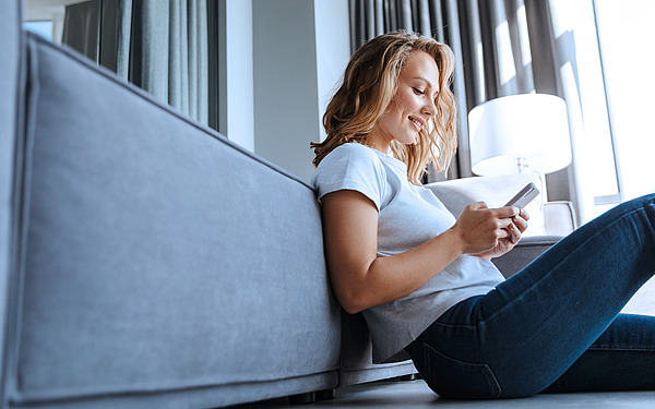 Eine Frau sitzt auf dem Boden, lehnt an einem grauen Sofa und lächelt, während sie auf ihr Handy schaut. Sie trägt ein weißes T-Shirt und Jeans. Helles Tageslicht fällt durch die großen Fenster hinter ihr.