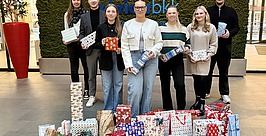 Sieben Personen stehen in einem Raum vor einer grünen Wand mit Firmenlogo und halten eingepackte Geschenke in der Hand. Zahlreiche bunt verpackte Geschenke sind vor ihnen auf dem Boden gestapelt. Sie lächeln und posieren für das Foto.