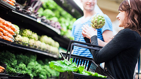 Eine Frau, die einen mit Salat gefüllten Einkaufskorb in der Hand hält, lächelt einen Mann an, während sie in der Gemüseabteilung eines Lebensmittelgeschäfts frisches Gemüse, darunter Artischocken, einkaufen.