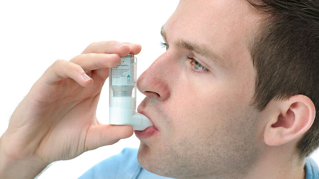 Eine Person benutzt einen Inhalator, hält ihn an den Mund und drückt auf den Behälter, um das Medikament zu inhalieren, vor einem weißen Hintergrund.