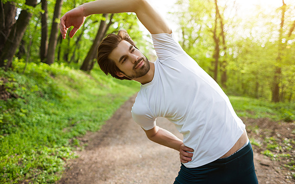 Ein Mann in einem weißen T-Shirt streckt seine Seite mit einem Arm über dem Kopf, während er auf einem Waldweg steht, umgeben von grünen Bäumen und Sonnenlicht.
