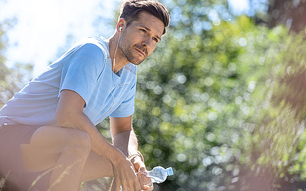 Ein Mann in hellblauem Hemd und Shorts, mit Kopfhörern, hockt im Freien und hält eine Wasserflasche in der Hand. Er wirkt nachdenklich und blickt in die Ferne. Im Hintergrund ist üppiges grünes Laub im hellen Sonnenlicht zu sehen.