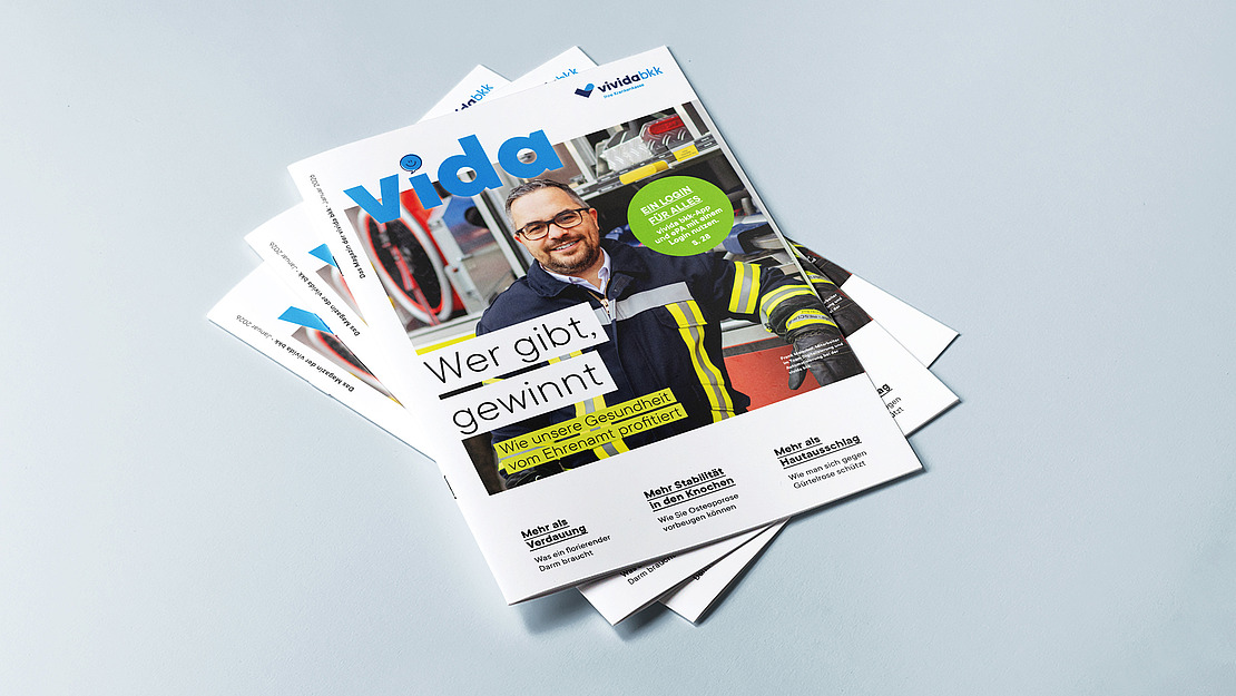 Ein Stapel "vida"-Magazine auf hellblauem Untergrund; die oberste Ausgabe zeigt einen lächelnden Mann im Feuerwehranzug und mit Brille, mit deutschem Text und einem grünen kreisförmigen Etikett auf dem Umschlag.