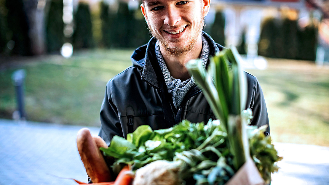 Ein junger Mann lächelt im Freien und hält eine Papiertüte mit frischem Gemüse in der Hand, darunter Frühlingszwiebeln, Karotten und Blattgemüse. Der Hintergrund ist unscharf und zeigt Bäume und eine gepflasterte Fläche.