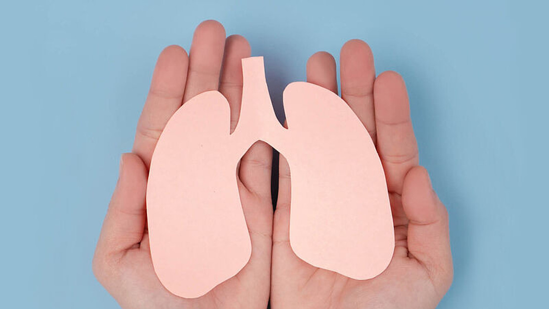 Zwei Hände halten sanft einen rosa Papierausschnitt einer menschlichen Lunge vor einem hellblauen Hintergrund, der die Gesundheit und Pflege der Lunge symbolisiert.