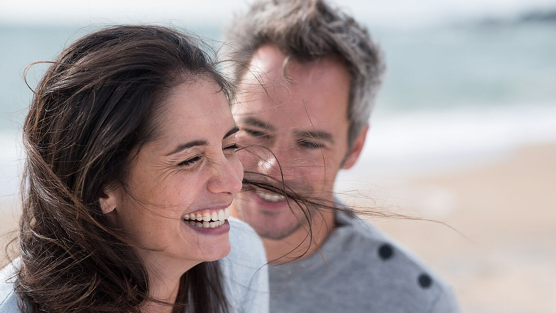 Eine lächelnde Frau mit dunklem Haar lacht, während ein Mann mit grauem Haar dicht hinter ihr am Strand steht. Beide wirken glücklich und entspannt. Der Hintergrund ist mit Sand und Meer verschwommen.