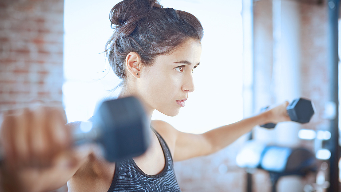 Eine Frau in Sportkleidung hält in jeder Hand eine Hantel, die Arme sind auf Schulterhöhe nach außen ausgestreckt, und trainiert konzentriert in einem gut beleuchteten Fitnessstudio oder Fitnessraum.
