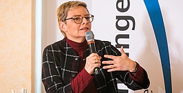 Eine Person mit kurzen blonden Haaren und Brille spricht in ein Mikrofon und gestikuliert dabei mit einer Hand. Sie trägt eine schwarz-weiß karierte Jacke über einem kastanienbraunen Rollkragenpullover. Auf dem Tisch stehen Bücher und Wassergläser.