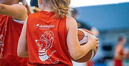 Ein junges Mädchen in einem roten Sporttrikot hält einen Basketball unter dem Arm. Sie dreht der Kamera den Rücken zu. Auf dem Trikot sind eine fröhliche Cartoon-Maus und das „vividabkk“-Logo aufgedruckt. Andere Spieler sind im Hintergrund unscharf zu sehen.