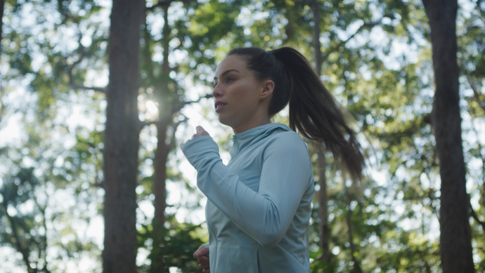 Eine Person joggt an einem sonnigen Tag durch ein Waldstück und trägt einen hellblauen Kapuzenpullover. Sie ist von hohen Bäumen mit grünen Blättern umgeben und das Sonnenlicht fällt durch die Zweige und schafft eine friedliche Atmosphäre.